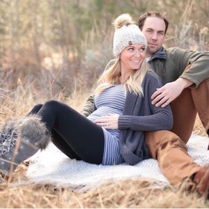 May include: A person wearing a beige knit beanie with a pom-pom, a grey striped long-sleeve shirt, a dark grey cardigan, black leggings, and grey winter boots with faux fur trim, sitting next to a person in an olive green jacket and brown trousers.