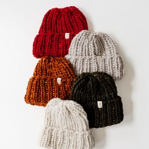 May include: A collection of five chunky knit beanies in red, light grey, burnt orange, dark green, and cream. Each hat features a thick ribbed cuff and a small rectangular tag with the text p&p attached to the front of the folded brim.