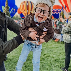 May include: A child wearing a beige knit beanie with a pom-pom, black-rimmed glasses, a brown button-up jacket, a light blue collared shirt, and light blue denim jeans. A small circular pin with text is attached to the jacket lapel.