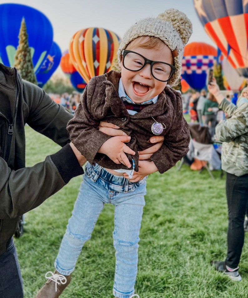 May include: A child wearing a beige knit beanie with a pom-pom, black-rimmed glasses, a brown button-up jacket, a light blue collared shirt, and light blue denim jeans. A small circular pin with text is attached to the jacket lapel.