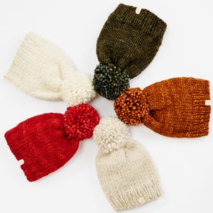 May include: Five chunky knit beanies arranged in a circular pattern. The hats are coloured cream, olive green, burnt orange, bright red, and beige. Each beanie features a large, matching or contrasting yarn pom-pom on top and a small tag.