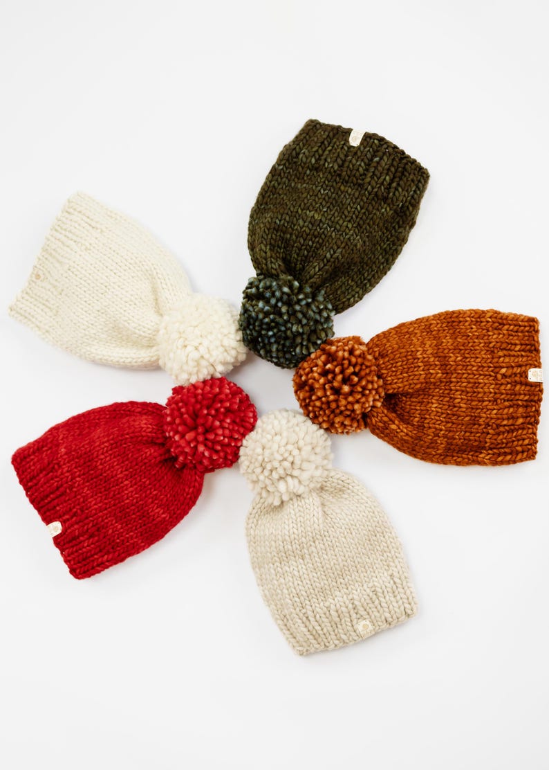 May include: Five chunky knit beanies arranged in a circular pattern. The hats are coloured cream, olive green, burnt orange, bright red, and beige. Each beanie features a large, matching or contrasting yarn pom-pom on top and a small tag.