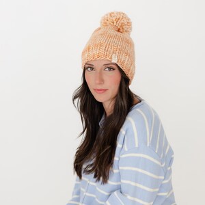 May include: A chunky knit beanie in a variegated peach and cream yarn, featuring a large matching pom-pom on top and a small rectangular brand tag on the folded brim. The hat is worn with a light blue and white horizontal striped jumper.