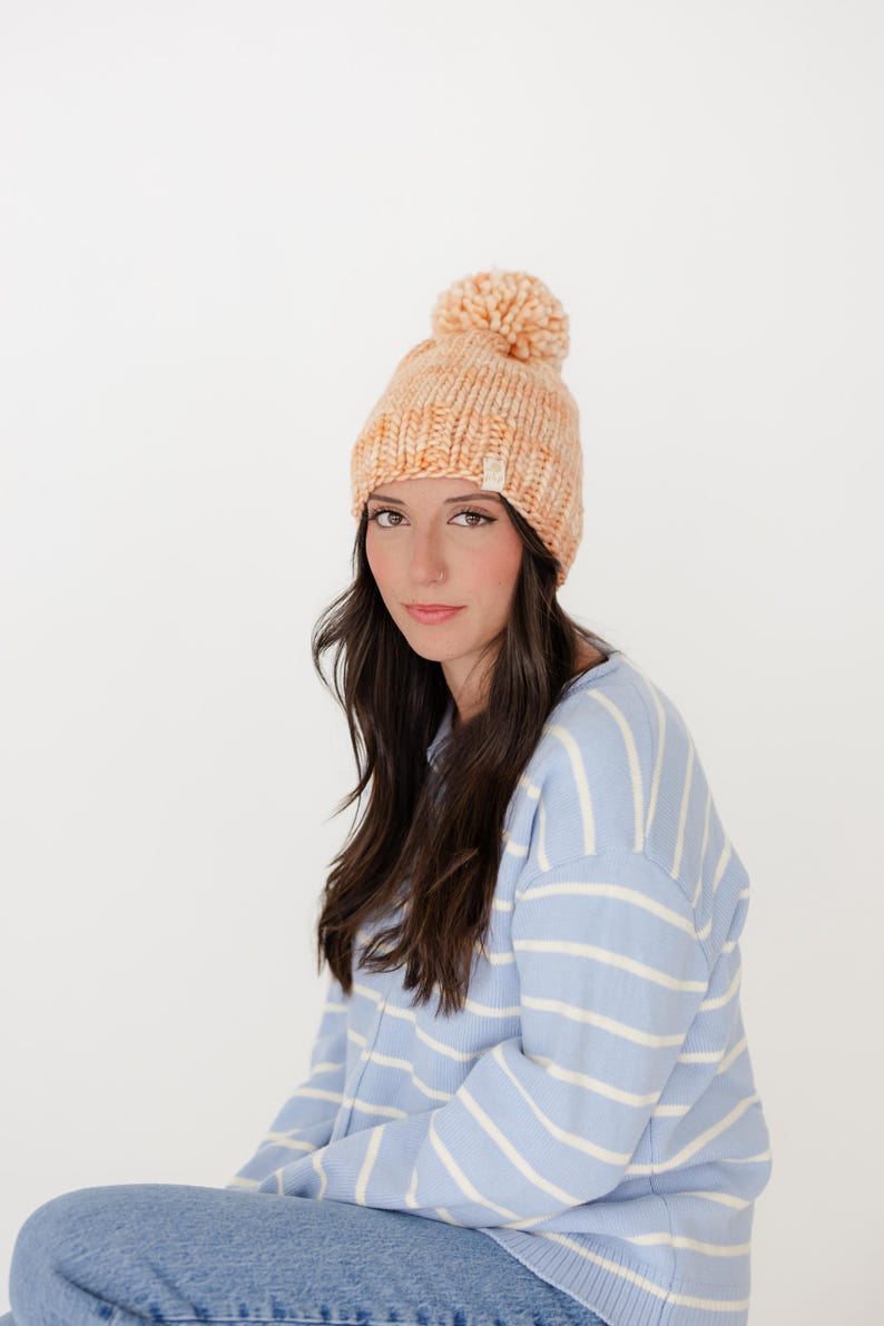 May include: A chunky knit beanie in a variegated peach and cream yarn, featuring a large matching pom-pom on top and a small rectangular brand tag on the folded brim. The hat is worn with a light blue and white horizontal striped jumper.
