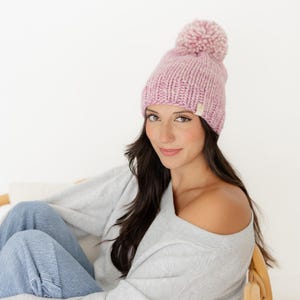 May include: A chunky knit beanie in a soft, variegated light pink yarn. The hat features a large, matching pom-pom on top and a small, rectangular beige fabric tag attached to the lower edge, showcasing a textured, hand-knit appearance.