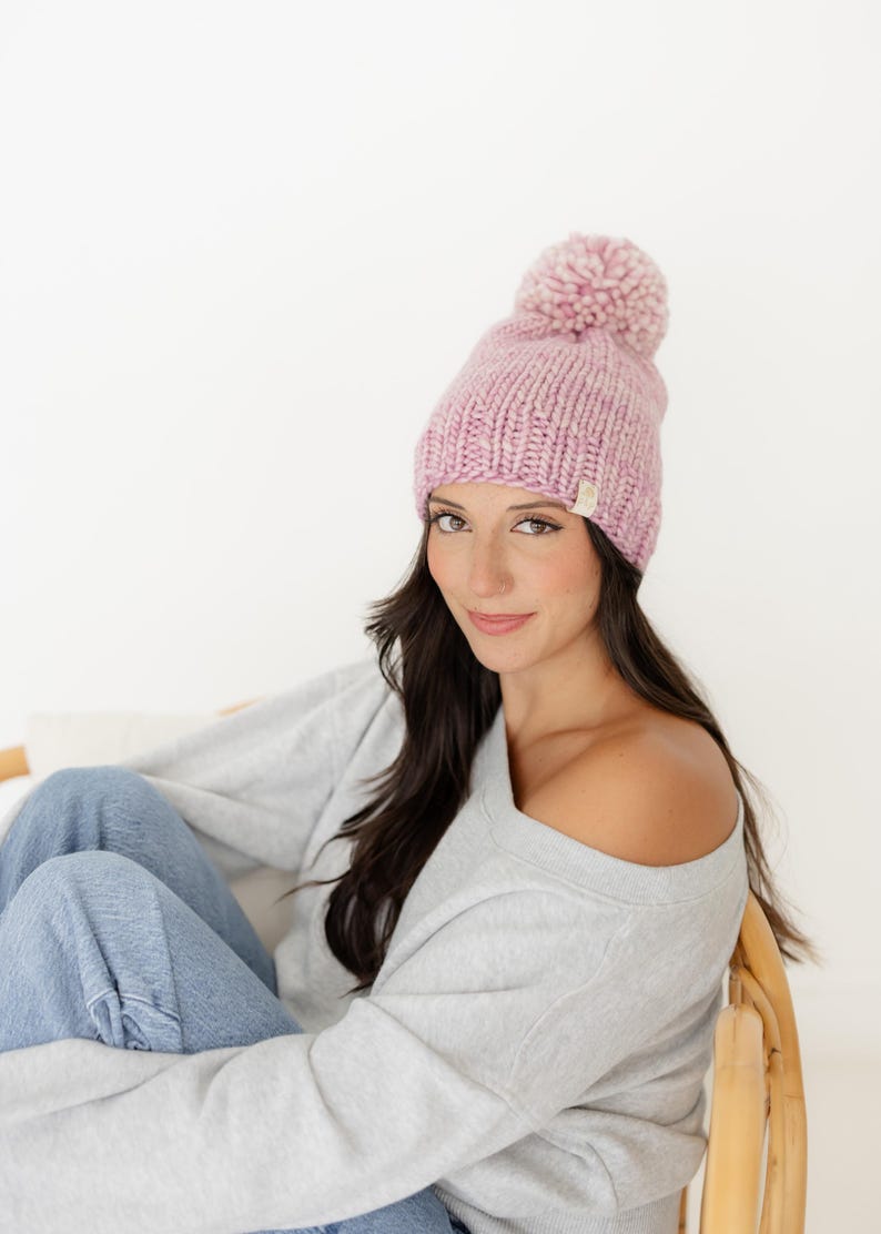 May include: A chunky knit beanie in a soft, variegated light pink yarn. The hat features a large, matching pom-pom on top and a small, rectangular beige fabric tag attached to the lower edge, showcasing a textured, hand-knit appearance.
