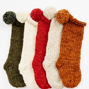 Könnte beinhalten: Fünf grob gestrickte Strümpfe nebeneinander, jeder mit einem großen Bommel an der Oberseite. Die Farben von links nach rechts sind Dunkelolivgrün, Creme, Hellrot, Cremeweiß und gebranntes Orange, gefertigt aus dickem Garn.