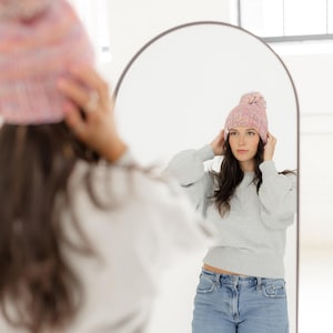 May include: A chunky knit beanie in a variegated pastel colour palette featuring shades of pink, blue, and yellow. The hat has a thick ribbed cuff and a large matching pom-pom on top, shown being worn in a reflection in an arched mirror.