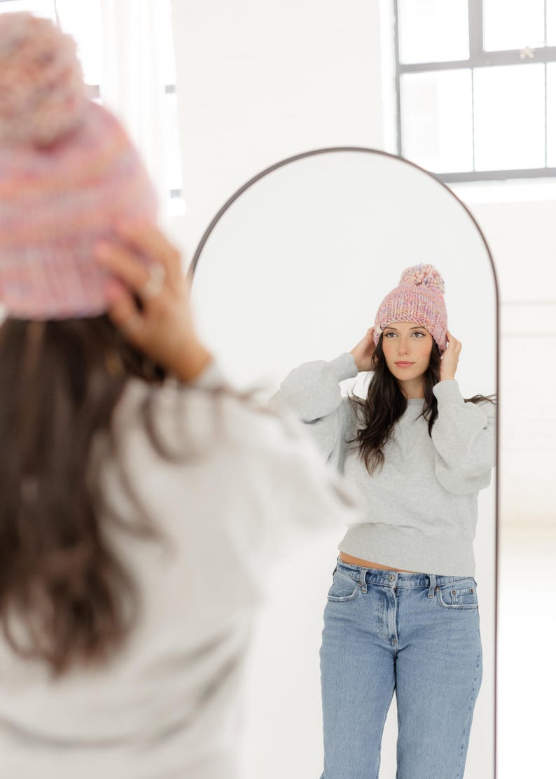May include: A chunky knit beanie in a variegated pastel colour palette featuring shades of pink, blue, and yellow. The hat has a thick ribbed cuff and a large matching pom-pom on top, shown being worn in a reflection in an arched mirror.