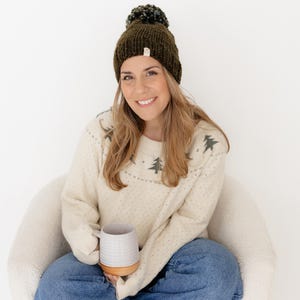 May include: A dark olive green knit beanie with a matching pom-pom on top, paired with a cream-coloured long-sleeve sweater featuring a repeating dark green pine tree pattern across the chest and shoulders, worn with blue denim jeans.