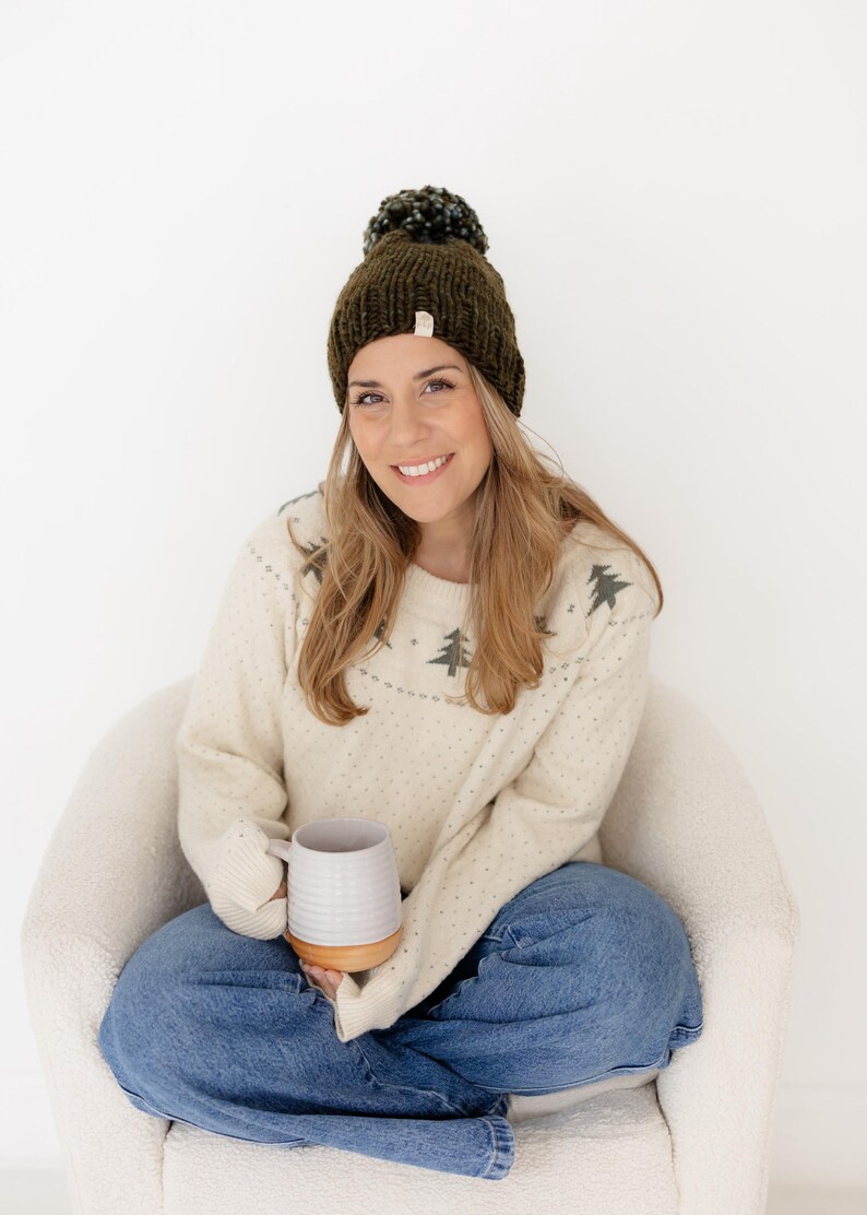 May include: A dark olive green knit beanie with a matching pom-pom on top, paired with a cream-coloured long-sleeve sweater featuring a repeating dark green pine tree pattern across the chest and shoulders, worn with blue denim jeans.