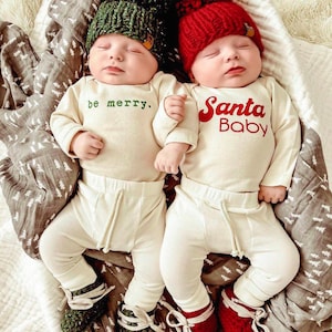 May include: Two infants wearing cream-coloured long-sleeve bodysuits and ribbed trousers. One bodysuit has the green text "be merry." and a green pom-pom hat. The other has the red text "Santa Baby" and a red pom-pom hat. Both wear matching knit booties.