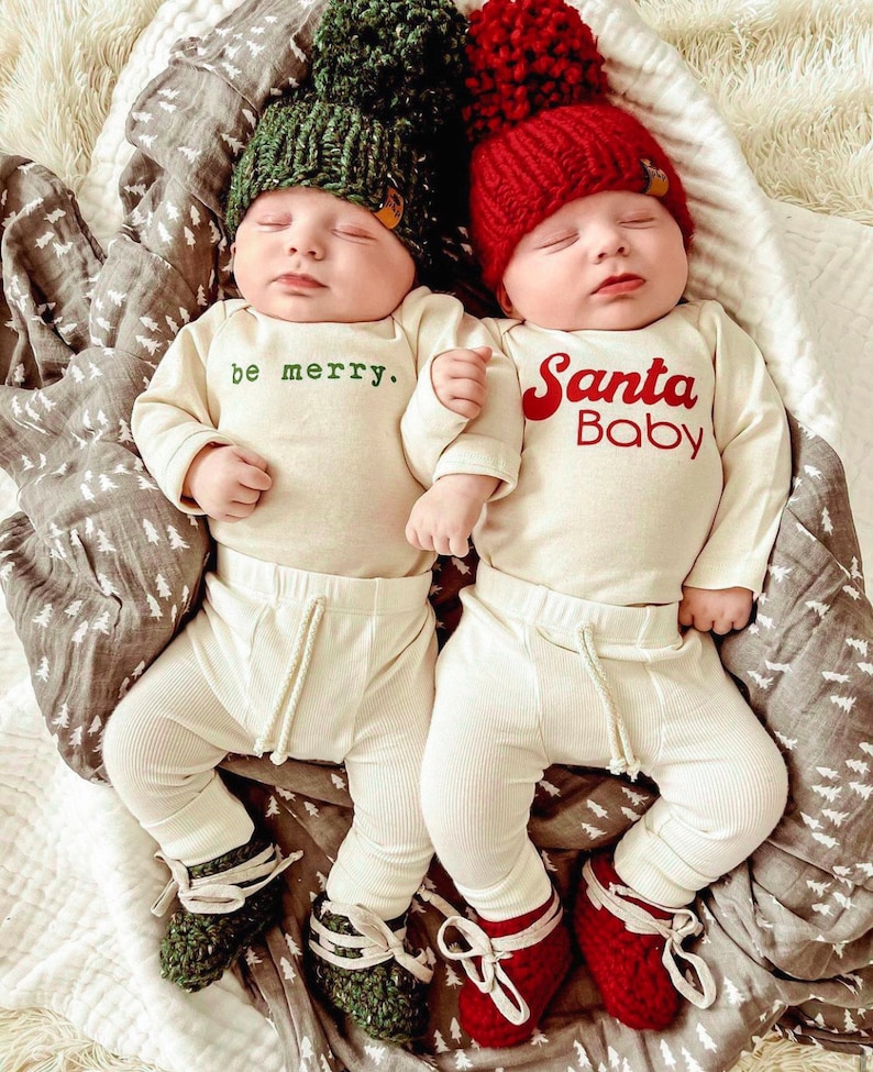 May include: Two infants wearing cream-coloured long-sleeve bodysuits and ribbed trousers. One bodysuit has the green text "be merry." and a green pom-pom hat. The other has the red text "Santa Baby" and a red pom-pom hat. Both wear matching knit booties.