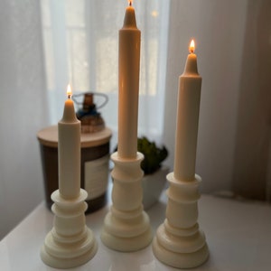 May include: Three white pillar candlesticks with white candles, each with a lit flame. The candlesticks are on a white surface.