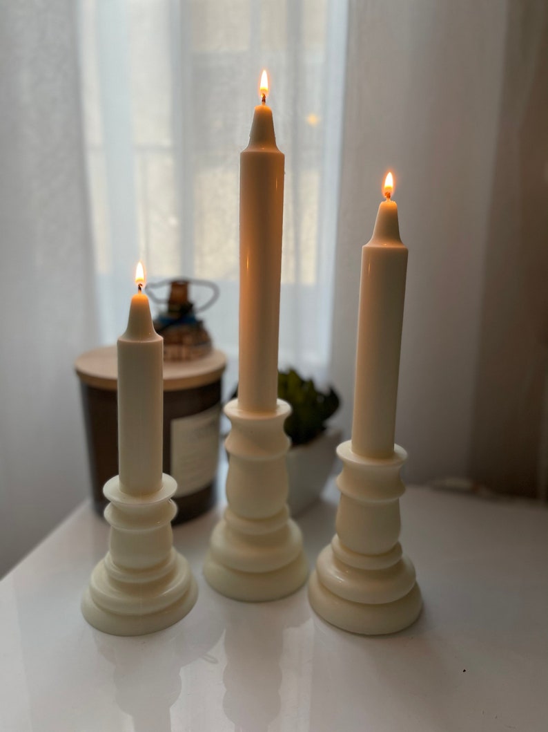 May include: Three white pillar candlesticks with white candles, each with a lit flame. The candlesticks are on a white surface.