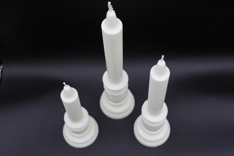 May include: Three white pillar candles of varying heights, each with a white wick. The candles are on a black surface.