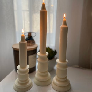 May include: Three white pillar candlesticks with white candles, two of which are lit. The candlesticks have a decorative, fluted design.