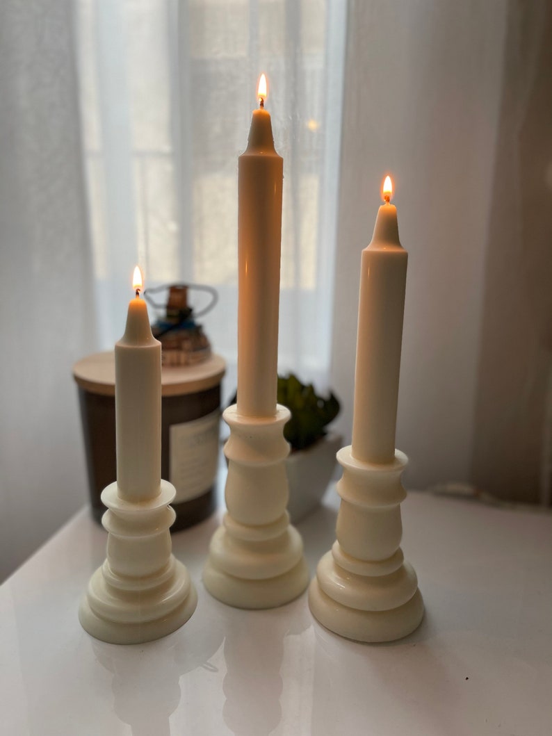 May include: Three white pillar candlesticks with white candles, two of which are lit. The candlesticks have a decorative, fluted design.