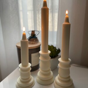 May include: Three white pillar candles with flames burning. The candles are on white candle holders with a decorative design. The candle holders are on a white surface.