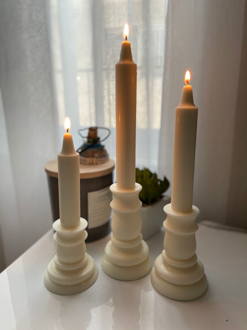 May include: Three white pillar candles with flames burning. The candles are on white candle holders with a decorative design. The candle holders are on a white surface.