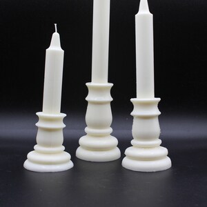 May include: Three white candle holders with white candles. The candle holders are made of ceramic and have a classic design.