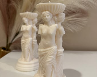 May include: Two white ceramic candle holders shaped like statues of women. The candle holders are on a white surface.