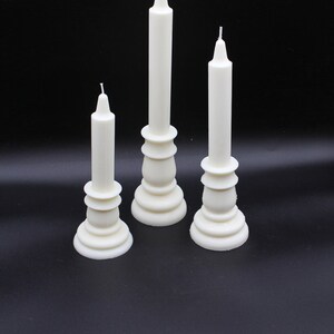 May include: Three white candlesticks with white candles on a black background. The candlesticks are different sizes and have a decorative design.