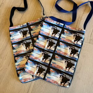 Könnte beinhalten: Zwei blaue und weiße Stofftaschen mit verstellbaren Trägern. Die Taschen haben ein sich wiederholendes Muster eines Berner Sennenhundes mit dem Text "REPE BRIQLE DE BAUROU" und "Bouvier Bernese", das auf jeder Tasche aufgedruckt ist.