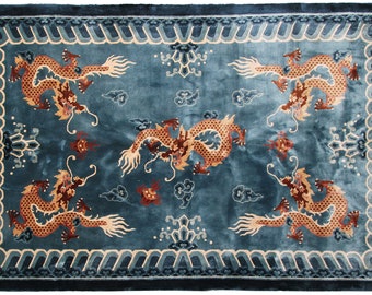 May include: Teal-colored rectangular rug with a detailed dragon design. The rug features four dragons in shades of brown and gold, along with cloud and floral motifs. The border has a wave-like pattern and fringe on the ends.