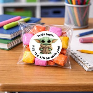 May include: A clear cellophane bag filled with colourful sweets, featuring a round sticker with a cartoon image of Grogu and the text "Yoda best! May the 4th be with you!" on a wooden desk with school supplies.