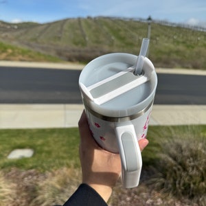 Pu&ograve; includere: Tazza da viaggio bianca con coperchio e manico grigi. Il coperchio ha una fessura per una cannuccia, e una cannuccia &egrave; inserita nella fessura. La tazza ha un design rosa e bianco con piccoli cuori rosa.