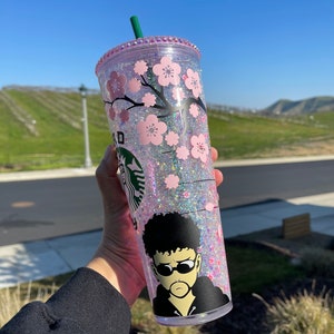 May include: A clear, glitter-filled tumbler with a pink lid and straw. The tumbler features a cherry blossom design and a graphic of a person wearing sunglasses. The Starbucks logo is visible.