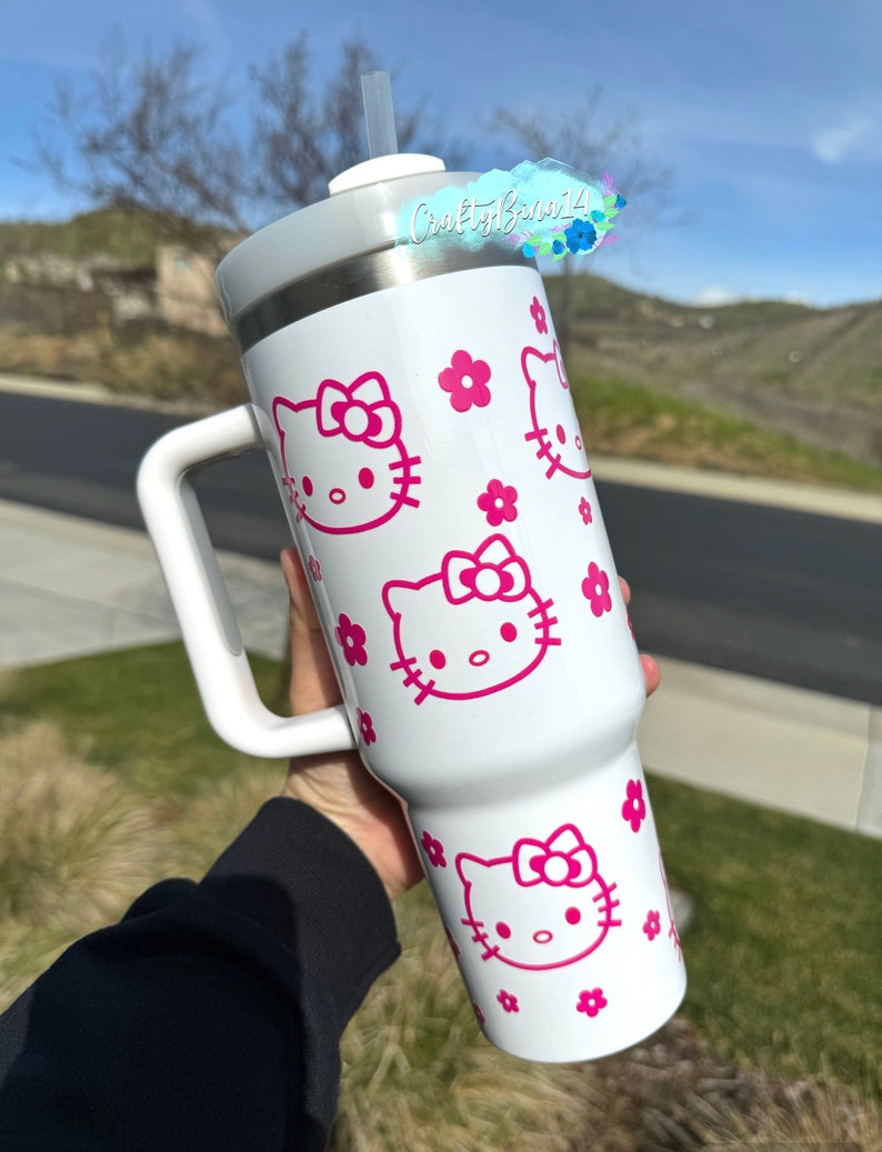 Pu&ograve; includere: Tazza da viaggio in acciaio inossidabile bianca con un design rosa Hello Kitty e un manico. La tazza ha un coperchio con una cannuccia ed &egrave; decorata con fiori rosa.