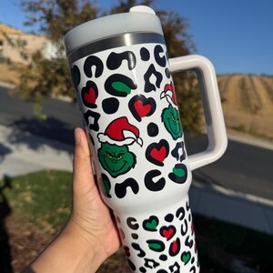 May include: White travel mug with a handle, featuring a Grinch and leopard print design. The design includes green Grinch faces wearing Santa hats, red hearts, and black and green leopard spots. The mug has a white handle and a silver rim.