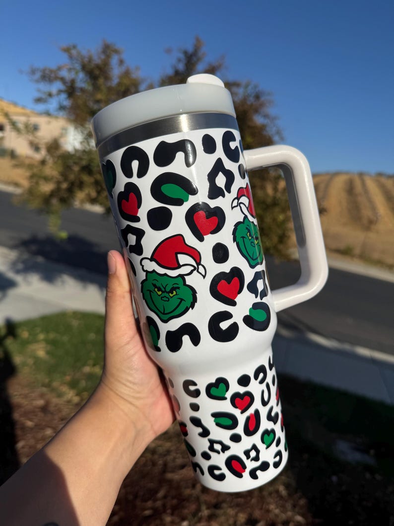 May include: White travel mug with a handle, featuring a Grinch and leopard print design. The design includes green Grinch faces wearing Santa hats, red hearts, and black and green leopard spots. The mug has a white handle and a silver rim.