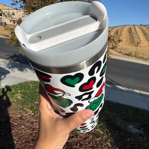 May include: A white tumbler with a lid, featuring a Grinch-themed design. The tumbler has a pattern of black spots, red hearts, and green Grinch faces. The lid is gray and has a white slider.