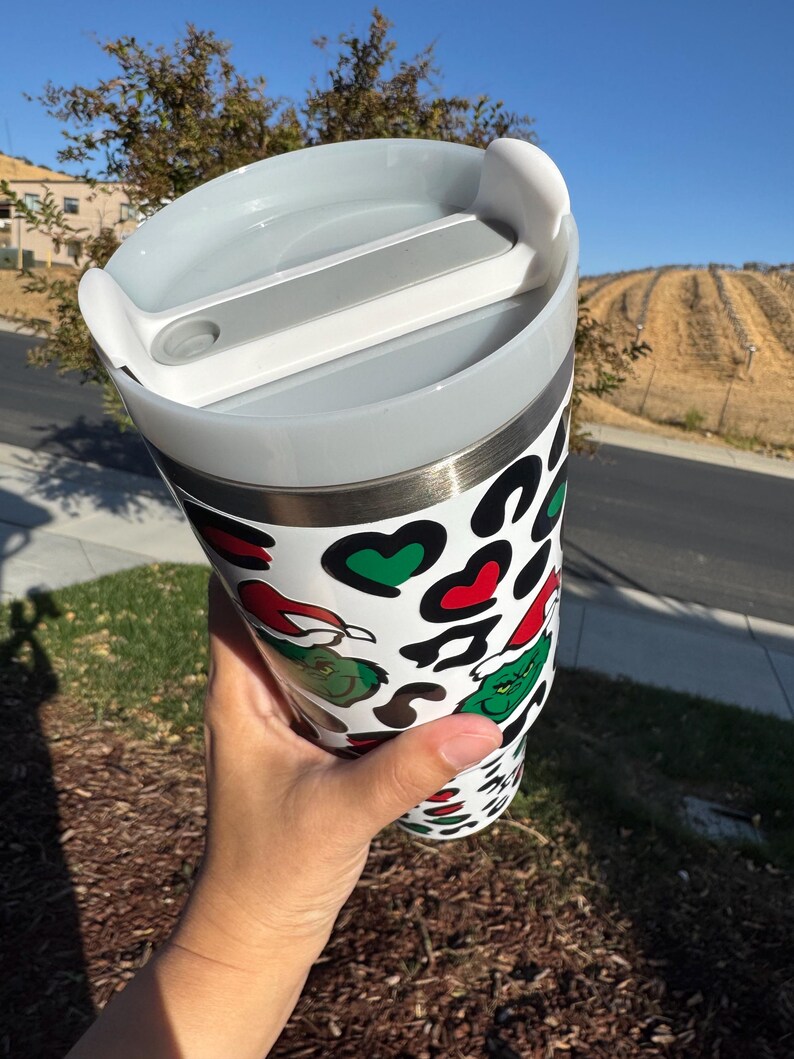 May include: A white tumbler with a lid, featuring a Grinch-themed design. The tumbler has a pattern of black spots, red hearts, and green Grinch faces. The lid is gray and has a white slider.