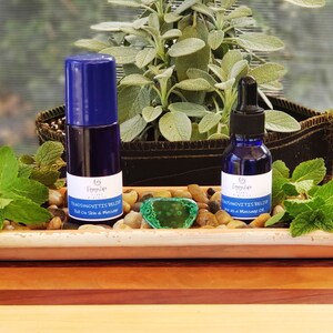 Puede incluir: Dos botellas de vidrio azul con etiquetas que dicen "TENSOMYOTITIS RELIEF" y "Roll On Skin & Massage Oil". Las botellas están sobre una bandeja de terracota con piedras verdes y una piedra verde en el centro. Hay hojas verdes en el fondo.