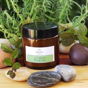 Puede incluir: Un tarro de vidrio marrón con tapa negra que contiene Cool Angelica Massage Salve. El tarro está sobre una superficie de madera con plantas verdes y piedras.