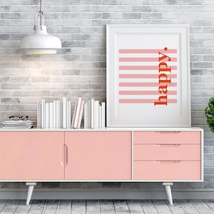 Könnte beinhalten: Eine rosa Kommode mit drei Schubladen und zwei Türen. Die Kommode hat weiße Beine und steht vor einer weißen Backsteinwand. Eine weiße Lampe hängt über der Kommode. Ein gerahmtes Bild mit rosa und weißen Streifen und dem Wort "happy" in roten Buchstaben hängt an der Wand über der Kommode. Eine kleine grüne Topfpflanze steht rechts neben der Kommode.