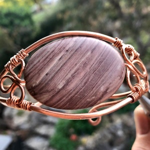 K&ouml;nnte beinhalten: Ein Armband aus Kupferdraht mit einem gro&szlig;en, ovalen, braun-wei&szlig; gestreiften Stein.