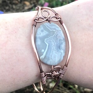 K&ouml;nnte beinhalten: Ein mit Kupferdraht umwickeltes Armband mit einem grauen und wei&szlig;en Achat-Stein. Das Armband besteht aus zwei Kupferdr&auml;hten, die miteinander verdreht sind, um einen sicheren Verschluss zu bilden.