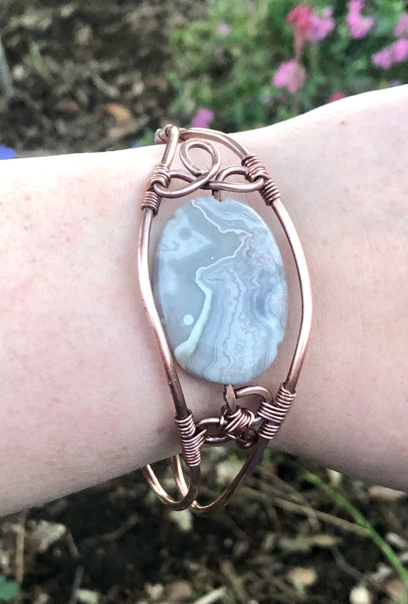 K&ouml;nnte beinhalten: Ein mit Kupferdraht umwickeltes Armband mit einem grauen und wei&szlig;en Achat-Stein. Das Armband besteht aus zwei Kupferdr&auml;hten, die miteinander verdreht sind, um einen sicheren Verschluss zu bilden.