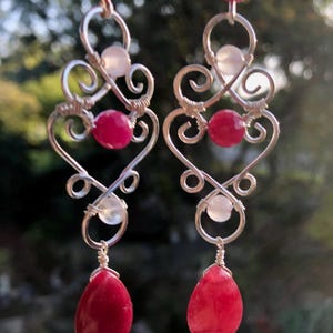 May include: A pair of handcrafted earrings with a silver wire design incorporating heart shapes. The earrings feature bright red and white beads, with a teardrop-shaped red bead at the base. The background is out of focus.