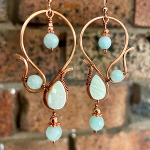 May include: Handmade copper earrings. Each earring has a distinctive shape, incorporating light blue beads and a pear-shaped, iridescent white stone. The design is intricate, with a handcrafted feel, and is suitable for everyday wear.