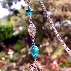 May include: Silver bookmark with a teal blue glass bead and a silver angel charm. The bookmark is decorated with a swirling pattern.