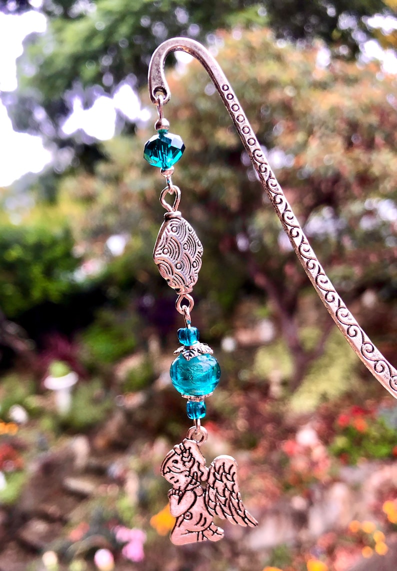 May include: Silver bookmark with a teal blue glass bead and a silver angel charm. The bookmark is decorated with a swirling pattern.