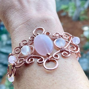 May include: A copper wire-wrapped bracelet featuring a large oval light pink gemstone at the centre, accented by smaller round translucent pink beads and intricate metal swirls. The design is constructed with coiled copper wire elements.