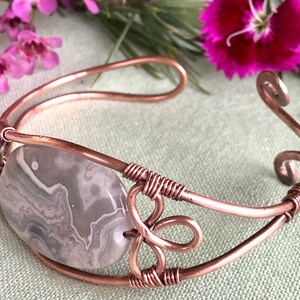 K&ouml;nnte beinhalten: Ein Armband aus Kupferdraht mit einem grauen und wei&szlig;en Achat-Stein. Das Armband hat ein zartes, filigranes Design mit einem Spiralmuster.