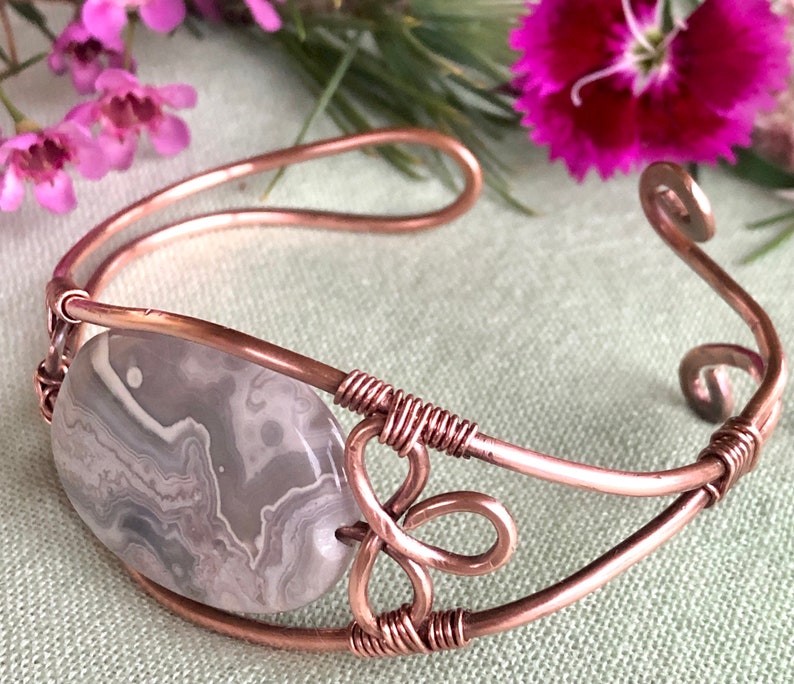 K&ouml;nnte beinhalten: Ein Armband aus Kupferdraht mit einem grauen und wei&szlig;en Achat-Stein. Das Armband hat ein zartes, filigranes Design mit einem Spiralmuster.